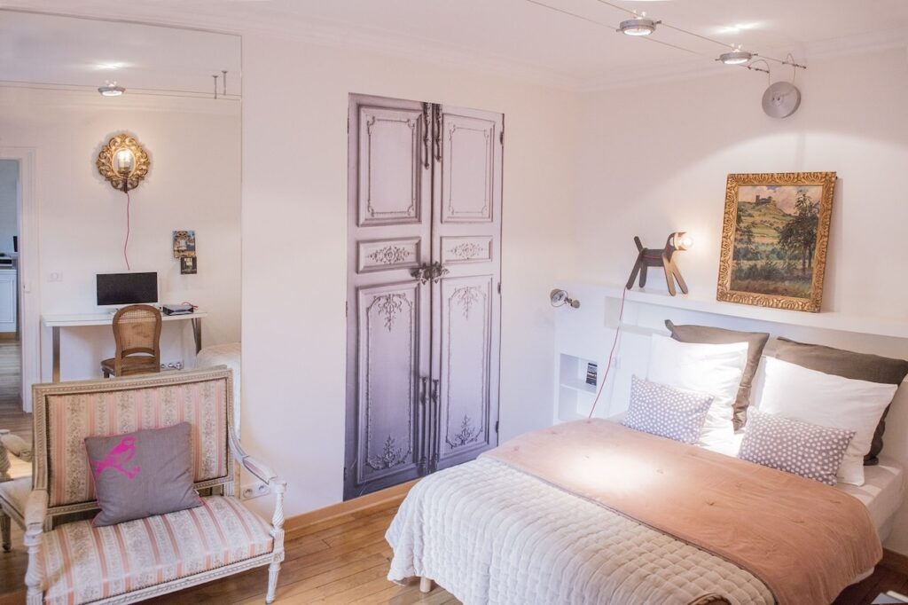 airbnbs in paris: Cozy bedroom interior featuring a white double bed with a beige quilt and multiple pillows, vintage-style upholstered armchair with a pink cushion, an ornate golden mirror, and whimsical wall art including a trompe-l'oeil painted door and a framed landscape painting. The room has a warm wooden floor and a small desk with a chair in the corner.