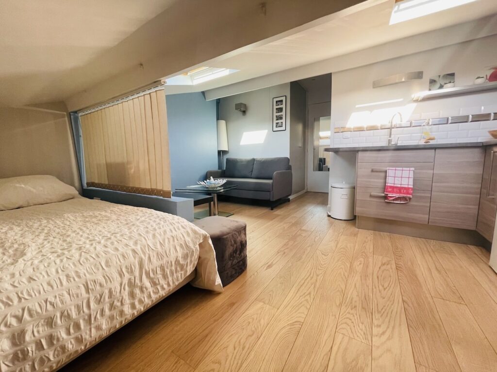 airbnbs in paris: Spacious studio apartment with ample natural light, featuring a comfortable double bed with a textured beige cover, a sleek kitchenette with modern fittings, and a cozy lounge area with a gray sofa and coffee table. The room is finished with warm wood flooring and a serene blue accent wall.