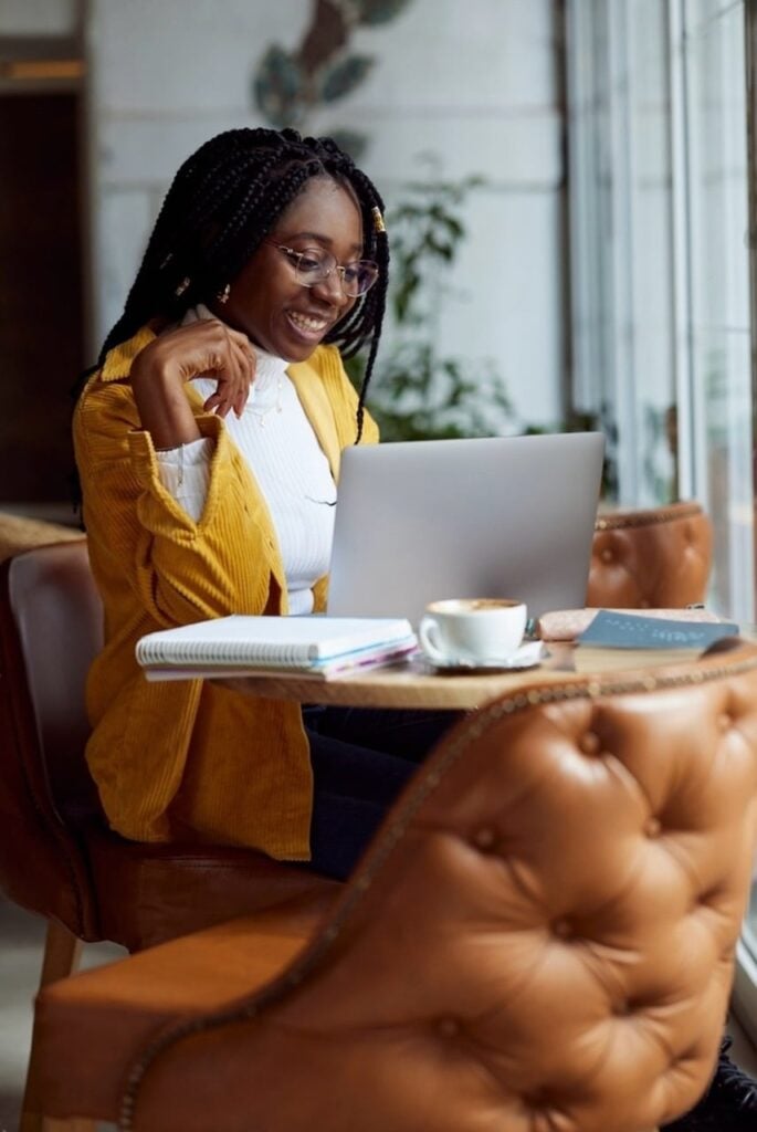 A joyful woman with braided hair, wearing glasses and a yellow cardigan, is working on a laptop in a cozy cafe setting. On her table, there's a cup of coffee, a notebook, and some colorful pens, enhancing the pleasant and productive atmosphere.