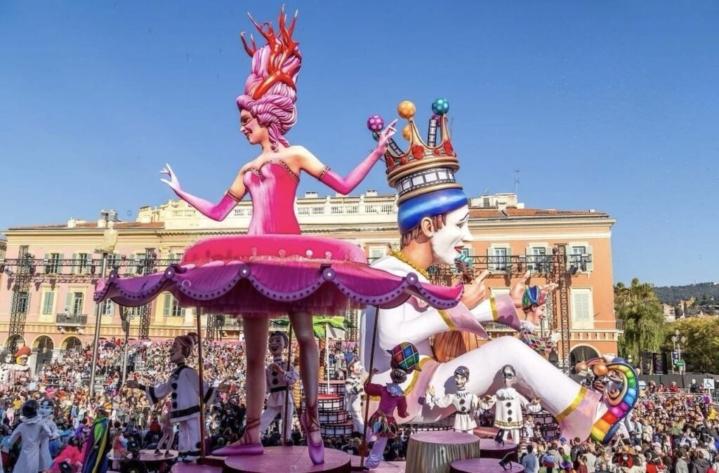 Vibrant and colorful float at the Carnival of Nice featuring a large pink-dressed figurine with an elaborate headpiece, accompanied by a jester playing drums, amidst a lively crowd of spectators under a clear blue sky.