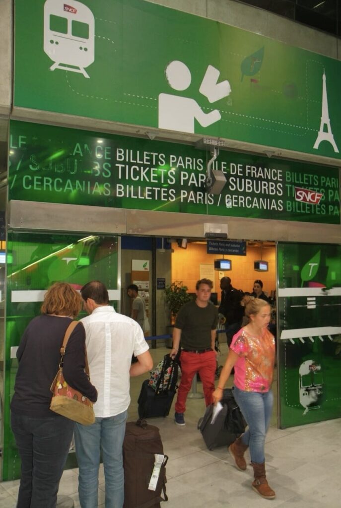Travelers queuing at a ticket counter with 'Billets Paris Ile de France' signage overhead, indicating a train ticket purchase point for local and regional travel in Paris. The green-themed decor features pictograms of a train and a person receiving a ticket, along with the iconic Eiffel Tower, conveying a busy transport hub atmosphere.
