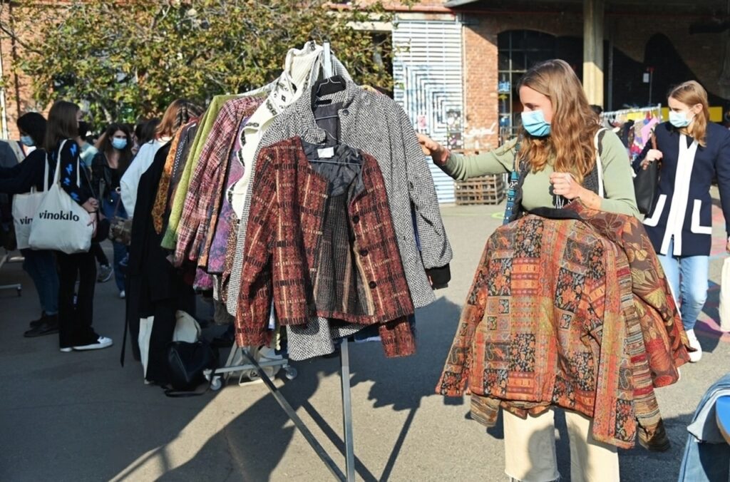 Vintage Shops in Paris: A woman in a mask examines a selection of vintage jackets on a clothing rack at an outdoor market, with other shoppers in the background adhering to health guidelines by wearing masks, highlighting a cautious return to normalcy in public spaces.