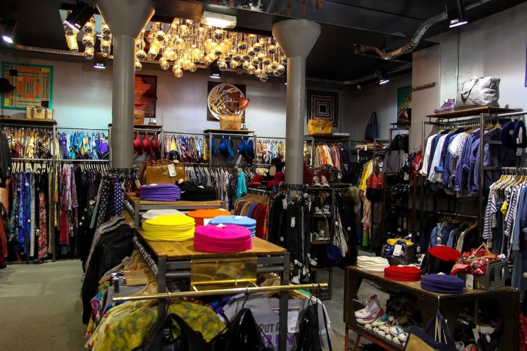 Vintage Shops in Paris: Interior of Kiliwatch, a chic vintage store with an array of colorful clothing on racks, vibrant berets on display, and an assortment of shoes below. The unique lighting fixture made of multiple bulbs adds a warm, retro ambiance to the shopping experience.