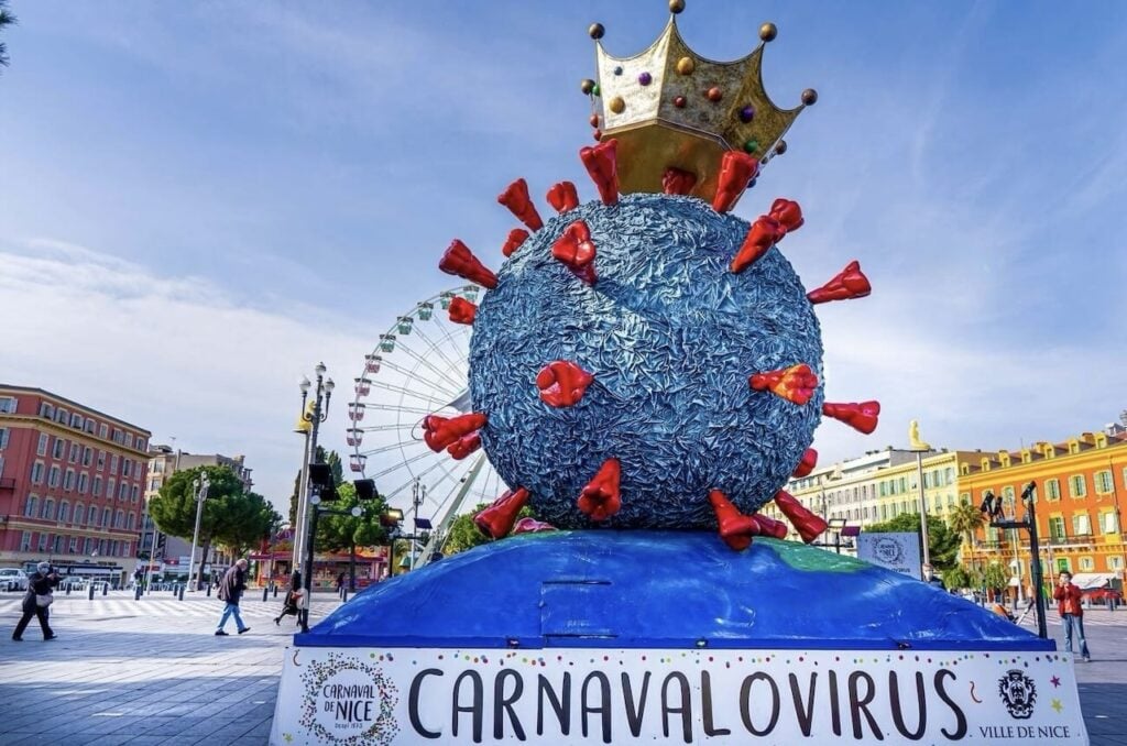 Carnaval de Nice: A satirical float at the Carnival of Nice featuring a giant blue sphere adorned with red spike proteins and a golden crown, humorously dubbed 'Carnavalovirus' against a backdrop of a Ferris wheel and the city's colorful buildings.