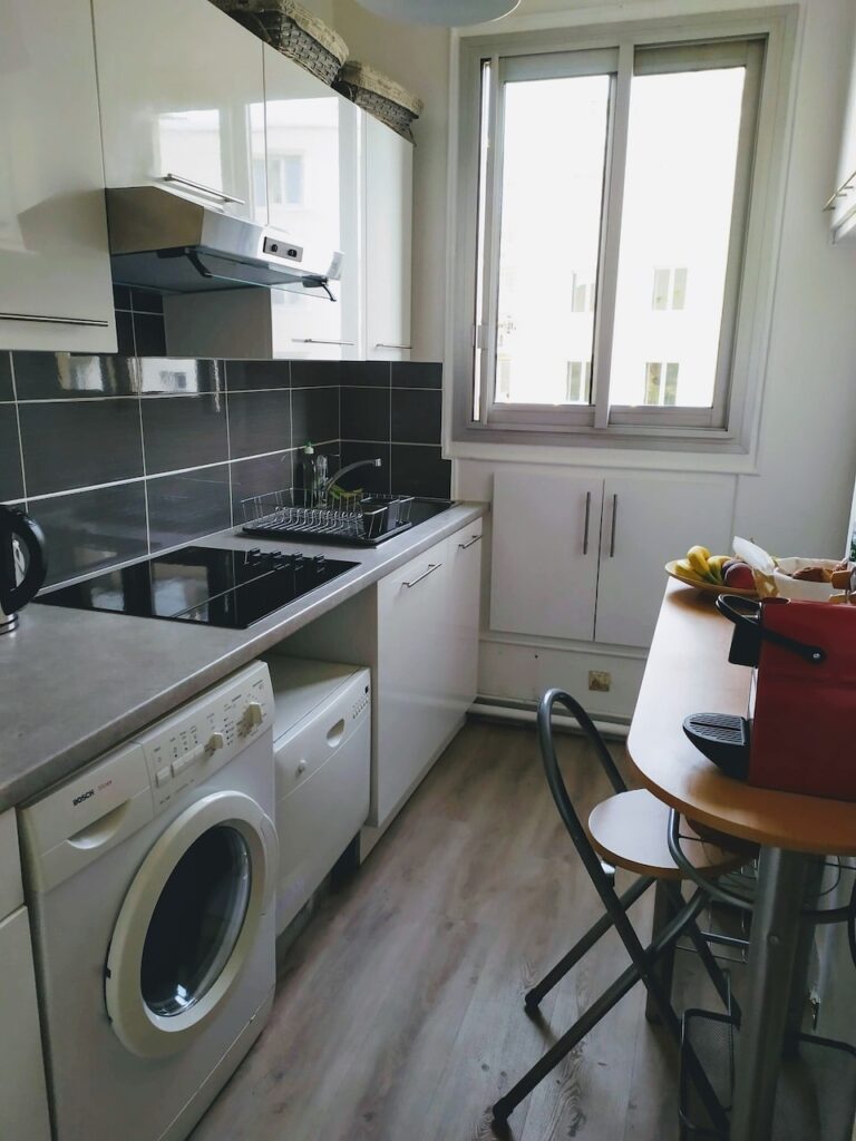 airbnbs in paris: Functional kitchen corner with white cabinets and dark countertops, equipped with a stainless steel stove and oven, an overhead exhaust hood, and a washing machine. A bright window lets in natural light, and a small dining table with two chairs offers a casual eating space, complemented by a basket of fresh fruit, adding a homey touch.