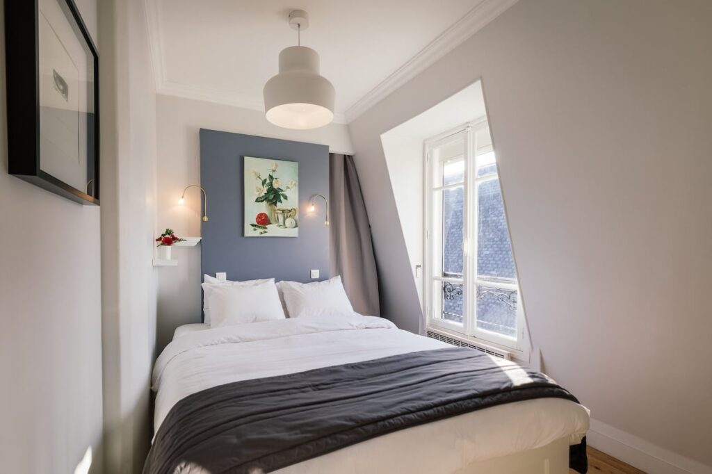 airbnbs in paris: Sleek and modern bedroom with a comfortable bed dressed in white linens and a dark throw, accented by a blue feature wall with floral artwork, elegant bedside lighting, and a view of the Parisian rooftops through an open window.
