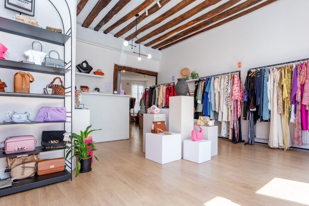 Vintage Shops in Paris: Modern and well-lit interior of Takk Paris, a boutique featuring an array of designer bags on sleek shelves and a selection of stylish clothing on racks, all under the rustic charm of exposed wooden ceiling beams.