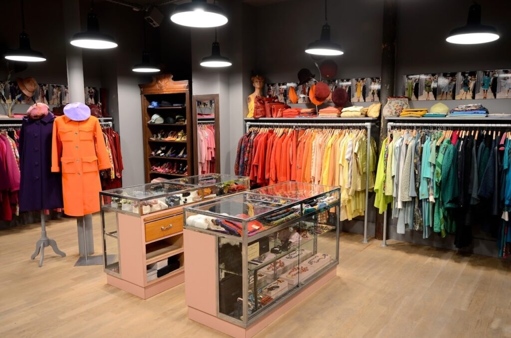 Vintage Shops in Paris: Spacious interior of 'Thanx God I'm a V.I.P.' vintage store, illuminated by pendant lights, featuring an elegant array of colorful clothing arranged by hue on racks, and a selection of accessories showcased in glass display cases.