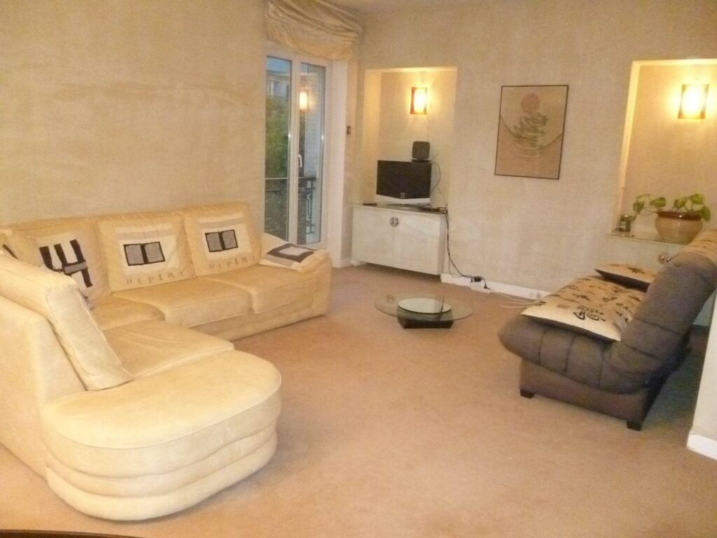 airbnbs in paris: A softly lit living room with plush beige sofas, decorative pillows with graphic designs, a modern glass coffee table, and a snug chaise lounge, all contributing to a serene and comfortable apartment setting.