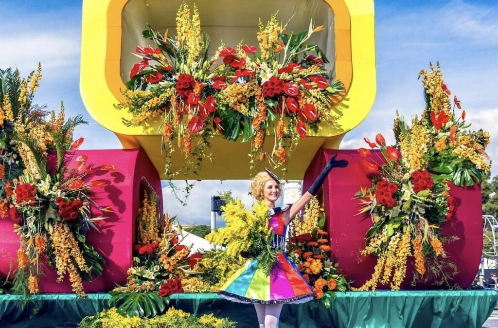 Carnaval de Nice: A woman in a vibrant, multicolored dress and black gloves poses gracefully on a float adorned with lush yellow and red flowers, part of a festive parade under a clear blue sky at the Carnival of Nice.