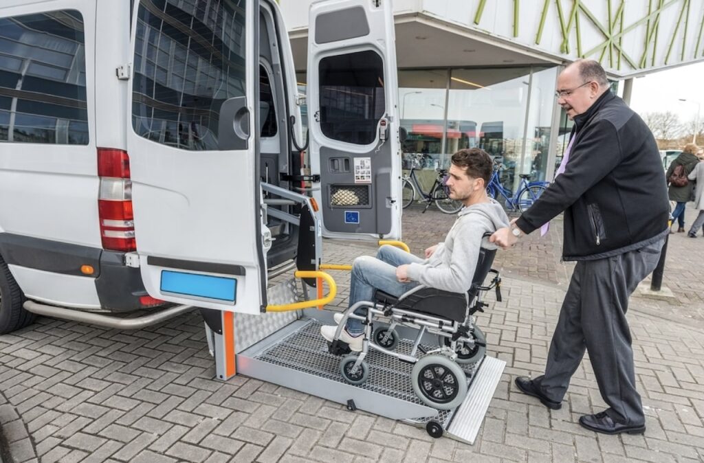 How to Get from CDG Airport to Paris: A taxi driver assists a man in a wheelchair onto a lift platform at the rear of an accessible van, suggesting a focus on transport for individuals with mobility challenges, outside a building with bicycles in the background.