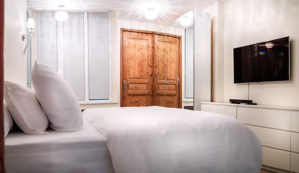 airbnbs in paris: Minimalist bedroom design with a comfortable bed set against a backdrop of natural wood wardrobe doors, white venetian blinds, and modern amenities including a wall-mounted flat-screen TV, combining contemporary style with classic touches.
