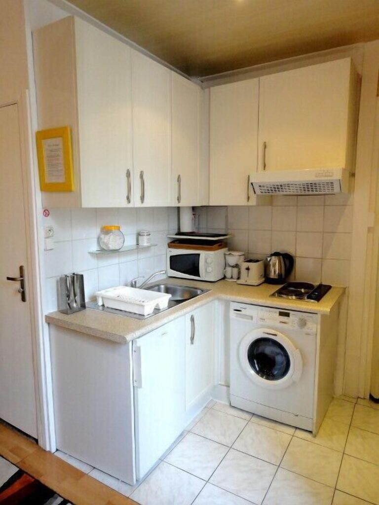 Compact and efficient kitchen space with white cabinetry, featuring a washing machine, an oven, a stainless-steel sink, and various kitchen appliances on the countertops.