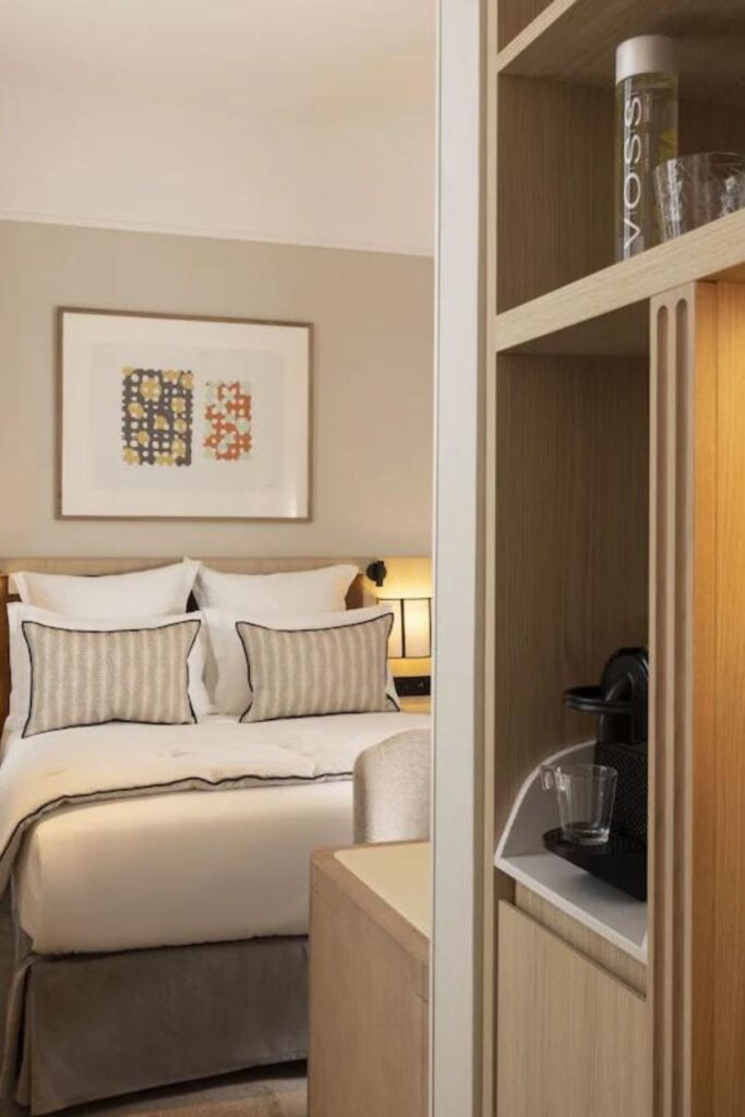 Elegant Paris hotel room featuring a plush double bed with striped pillows, ambient lighting, and artwork on the wall, as viewed from a connecting room that includes a shelf with a VOSS water bottle. Ideal for travelers searching for Paris hotels with connecting rooms.