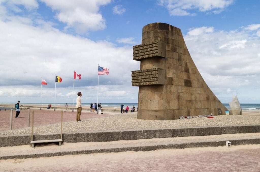 A young man gazes at the Omaha Beach memorial with national flags fluttering in the wind, representing a moment of history that can be experienced while on motorcycle tours in France.