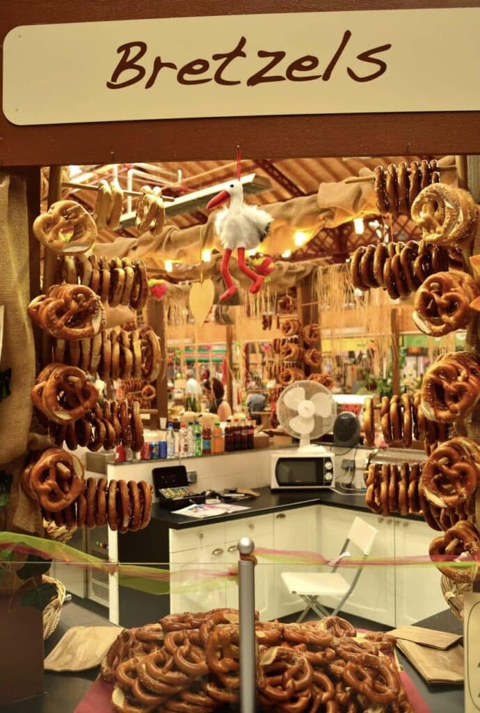 A stall selling traditional Alsatian bretzels at Marché Couvert de Colmar, featuring golden-brown pretzels hanging from rustic wooden displays. A small plush stork, a regional symbol of Alsace, adds a charming touch. A must-visit spot when spending one day in Colmar to enjoy authentic local flavors.