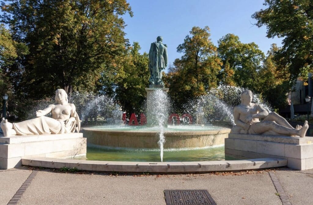 The elegant fountain at Parc du Champ de Mars in Colmar, France, featuring two reclining stone statues on either side and a bronze statue in the center. Water sprays beautifully from the fountain, surrounded by lush green trees under a clear blue sky. A serene spot to visit while exploring one day in Colmar.
