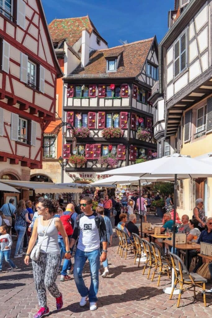 A lively scene in Grand Rue, the heart of Colmar’s Old Town, featuring colorful half-timbered buildings adorned with vibrant flowers. Locals and tourists stroll along the cobblestone streets, enjoying outdoor cafés and bustling market stalls. A must-visit spot when spending one day in Colmar to experience its charming Alsatian atmosphere.