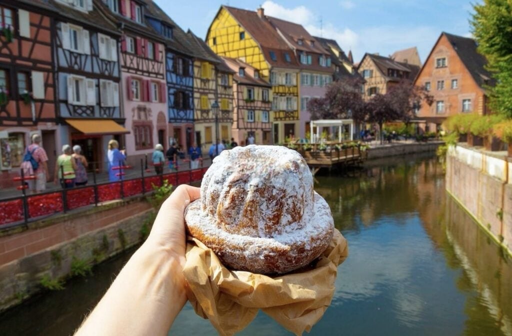 A freshly baked kougelhopf, a traditional Alsatian brioche dusted with powdered sugar, held in front of the colorful half-timbered houses of Little Venice in Colmar, France. The scenic canal and charming architecture make for a perfect backdrop while exploring one day in Colmar and savoring local delicacies.