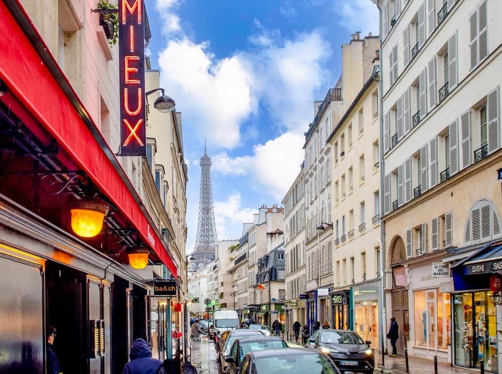 The charming streets of the best arrondissement to stay in Paris on a clear day, with the iconic Eiffel Tower in the distance and vibrant storefronts lining the road, as pedestrians and cars share the bustling thoroughfare.