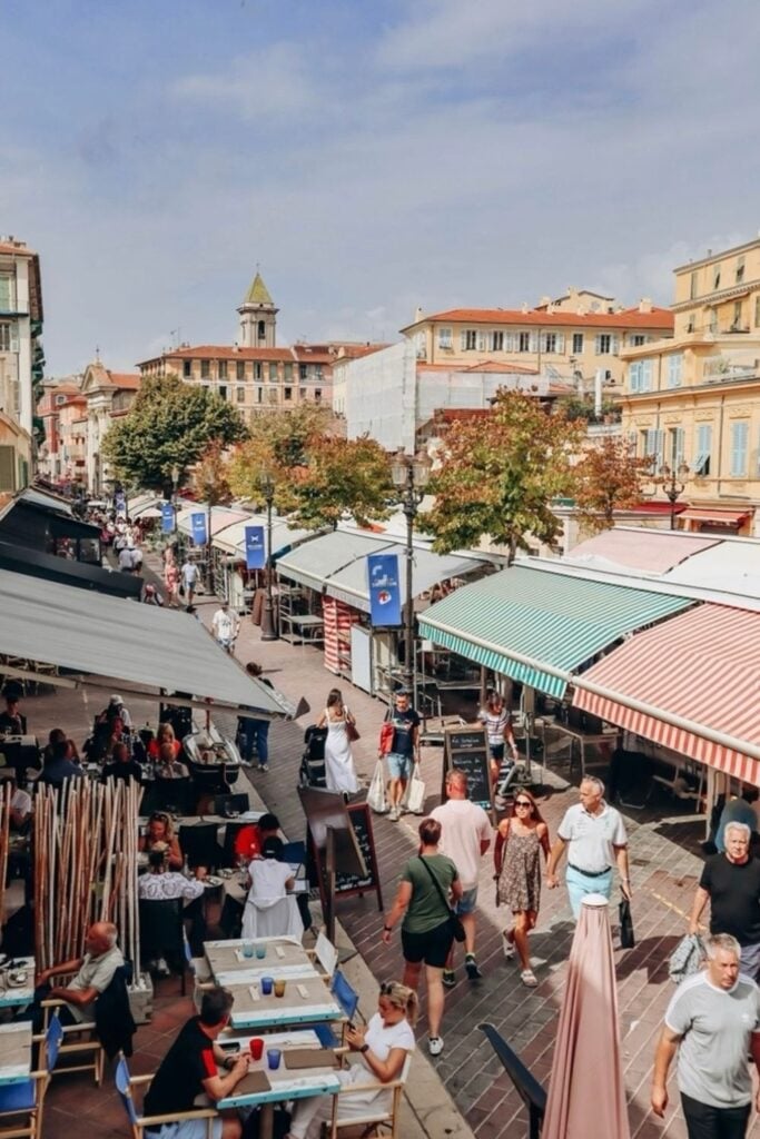 Casual outdoor dining and bustling street life on Cours Saleya in Nice, with pedestrians walking among the shaded market stalls and terraces. A scene that captures the lively urban rhythm for a Nice travel guide, highlighting the city's vibrant street culture and alfresco lifestyle.