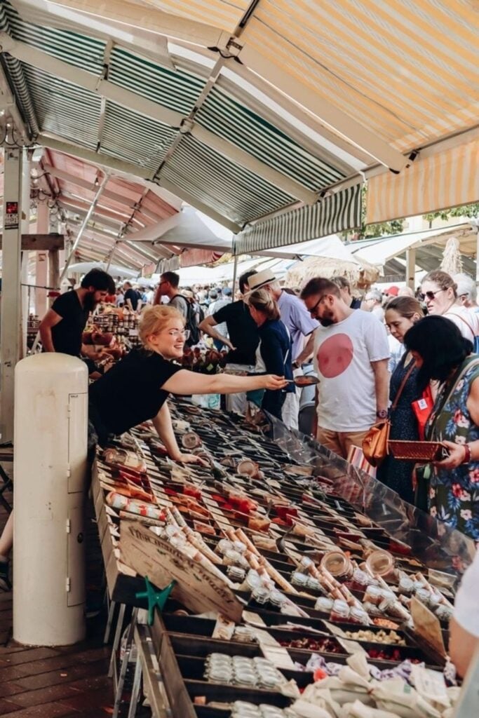 Bustling atmosphere at Cours Saleya market in Nice, with vendors and shoppers under striped awnings, engaging in the vibrant local shopping scene. This market is a must-visit for those exploring Nice, offering an authentic taste of the local culture and cuisine, ideal for inclusion in a Nice travel guide.