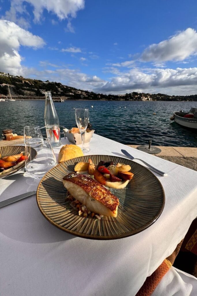 Enjoying gourmet dining at La Mère Germaine with a view of the serene Villefranche-sur-Mer bay. A plate with beautifully cooked fish and roasted vegetables is prominently displayed on a table set with wine glasses and a fresh baguette, embodying the culinary delights to be experienced while exploring things to do in Villefranche-sur-Mer.