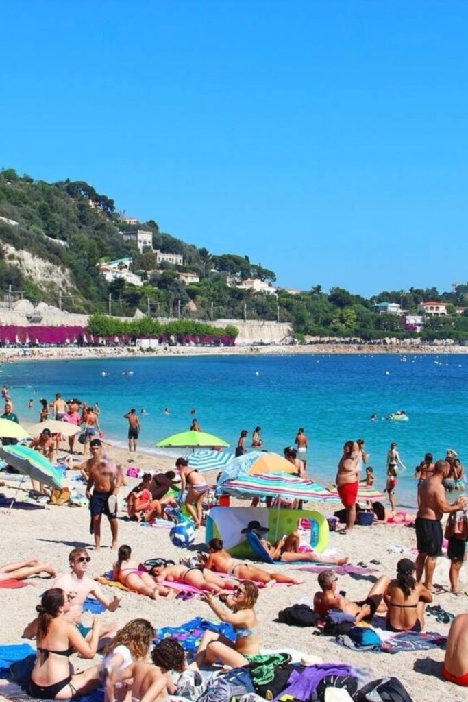 Vibrant beach day at Plage des Marinières in Villefranche-sur-Mer, with a crowd of beachgoers enjoying the sun on colorful towels and under parasols, set against a backdrop of the azure sea and lush hillside. The lively atmosphere is perfect for soaking in the sun and sea, a popular thing to do in Villefranche-sur-Mer.