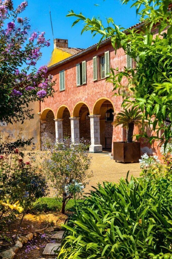 the peaceful courtyard of Saint Elme Citadel in Villefranche-sur-Mer, framed by an archway, with a lush garden in the foreground. The pink-hued walls and terracotta roof blend harmoniously with the surrounding greenery, offering a serene escape for those seeking historical things to do in Villefranche-sur-Mer.