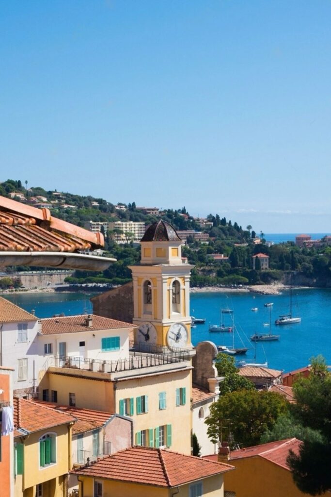 Breathtaking view from above Villefranche-sur-Mer, showcasing the historic Saint-Michel Church tower with its distinctive yellow façade and terracotta roof tiles, against the backdrop of the Mediterranean Sea with moored sailboats. This captivating vista is a highlight for visitors seeking picturesque things to do in Villefranche-sur-Mer.