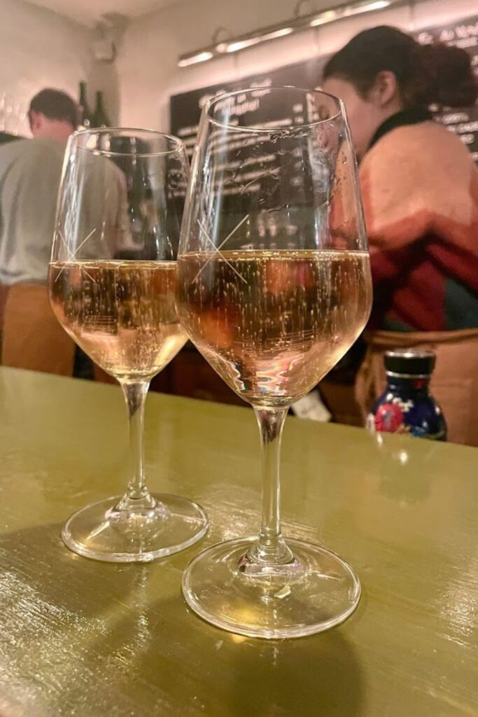Two glasses of rosé wine sit on a glossy gold counter, reflecting a lively bar scene, capturing the essence of Septime La Cave, celebrated for its curated wine selection among the best wine bars in Paris.
