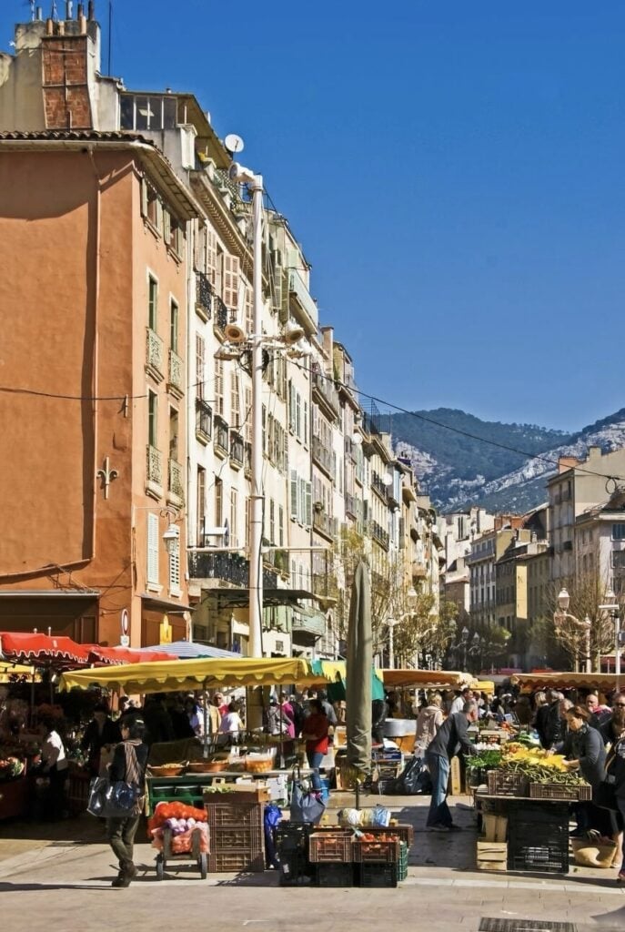 Warm-hued buildings with balconies adorned with flags line a sunlit street in Grasse, showcasing the city's vibrant charm. The historical architecture and clear blue skies make Grasse a picturesque, must-visit French Riviera cities.
