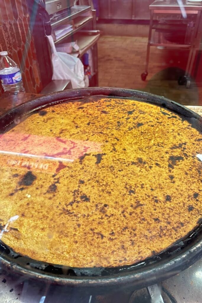 A large, freshly cooked Socca, the traditional chickpea flatbread, on a circular griddle in a Villefranche-sur-Mer eatery. The golden-brown surface and rustic kitchen setting invite food enthusiasts to taste this local delicacy, a flavorful addition to the culinary things to do in Villefranche-sur-Mer.