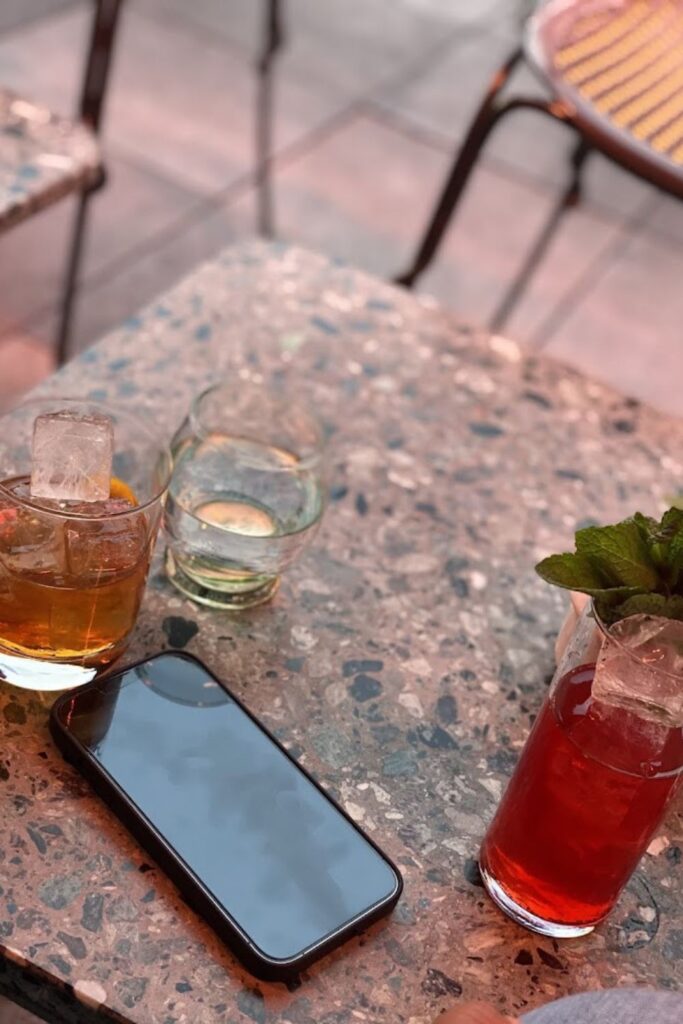 Alt text: A table at a rooftop bar with three different cocktails and a smartphone resting on it, surrounded by chairs. Best rooftop bar in Paris.