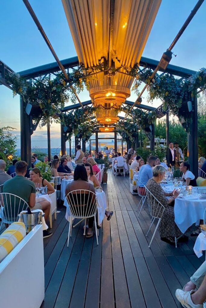 Alt text: People dining at a stylish rooftop bar with string lights and floral decorations, creating a cozy and elegant atmosphere as the sun sets. Best rooftop bar in Paris.