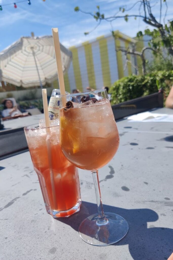 Alt text: Two colorful cocktails with bamboo straws sit on a table at a rooftop bar, with an umbrella and greenery in the background under a bright blue sky. Best rooftop bar in Paris.