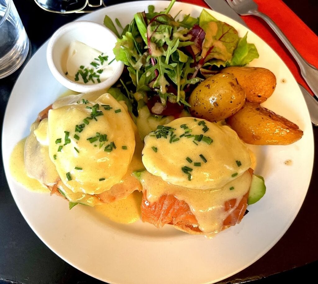 Elegant plating of Eggs Benedict at Eggs&Co. in Paris, featuring poached eggs on smoked salmon and English muffins, drizzled with creamy hollandaise sauce and garnished with chives. The dish is served with a side of roasted herbed potatoes and a fresh mixed green salad, embodying the best breakfast in Paris with a sophisticated flair.
