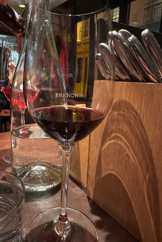 Close-up view of a red wine glass at Frenchie Bar à Vin, one of the cozy best wine bars in Paris. The wine glass, etched with the word 'FRENCHIE', is partially filled with red wine, with a background glimpse of the bar's warm, inviting interior and reflective windows showing the bustling Paris streets at night.
