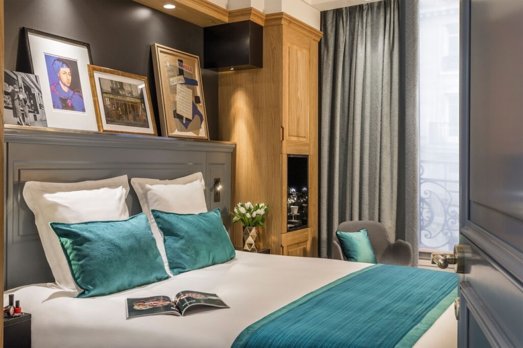Elegant room at Hôtel Ducs de Bourgogne in the 1st Arrondissement of Paris, featuring a comfortable bed with teal pillows and artwork above the headboard, showcasing stylish and cozy accommodation.