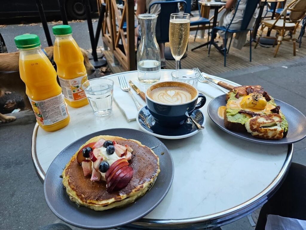 A delightful outdoor breakfast at Kozy Bosquet in Paris, complete with a stack of pancakes adorned with fresh fruit and whipped cream, an avocado toast topped with poached eggs and bacon, and a rich cappuccino with a heart-shaped design in the foam. The setting includes two bottles of orange juice and a glass of champagne, making it a luxurious contender for the best breakfast in Paris, enjoyed on a bustling street-side table.