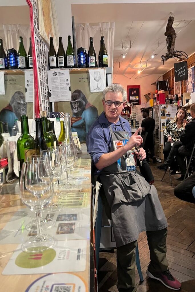 An animated scene inside best wine bars in Paris, showcasing a man in glasses and an apron standing behind a bar filled with empty wine bottles and glasses, ready for service. The bar's vibrant and eclectic décor, including a hanging sculpture and artwork of a gorilla, adds to the unique atmosphere of this Parisian wine bar, a go-to spot for wine enthusiasts.