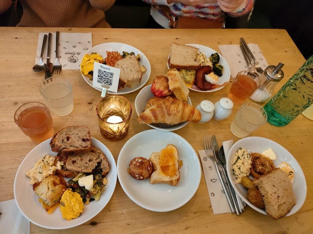 A bustling brunch table at La Bossue in Paris, laden with an array of dishes including scrambled eggs, assorted pastries like croissants and pain au chocolat, slices of rustic bread, and a colorful medley of vegetable sides. The table also features glasses of apple cider and a candle, creating a cozy atmosphere that exemplifies why this is considered one of the best breakfasts in Paris.