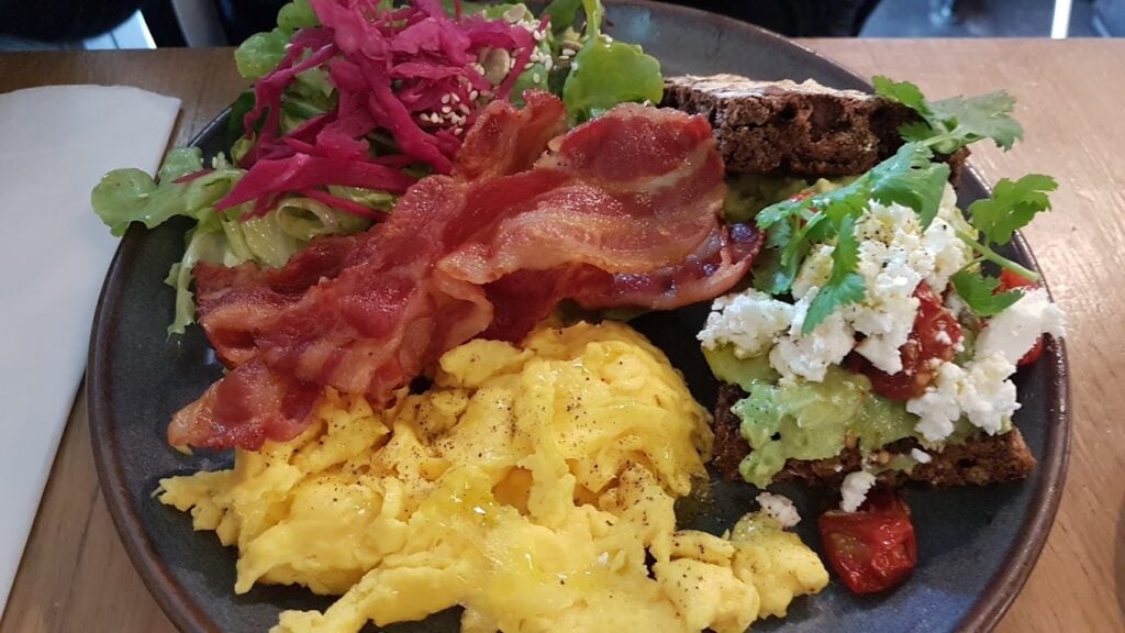 A vibrant plate from Le Bal Café in Paris, featuring fluffy scrambled eggs, crispy strips of bacon, and a slice of dark rye bread topped with avocado, cherry tomatoes, and crumbled feta cheese. The dish is garnished with a fresh green salad with a tangy dressing, presenting a balanced and visually appealing option for the best breakfast in Paris.