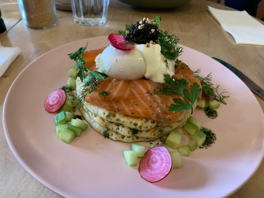Exquisite gourmet breakfast at The Hardware Société in Paris, featuring a perfectly stacked dish of herbed pancakes, smoked salmon, a poached egg topped with caviar, and garnished with fresh dill, parsley, and thinly sliced radishes. The combination of vibrant colors and refined ingredients on a soft pink plate showcases why this is celebrated as one of the best breakfasts in Paris.