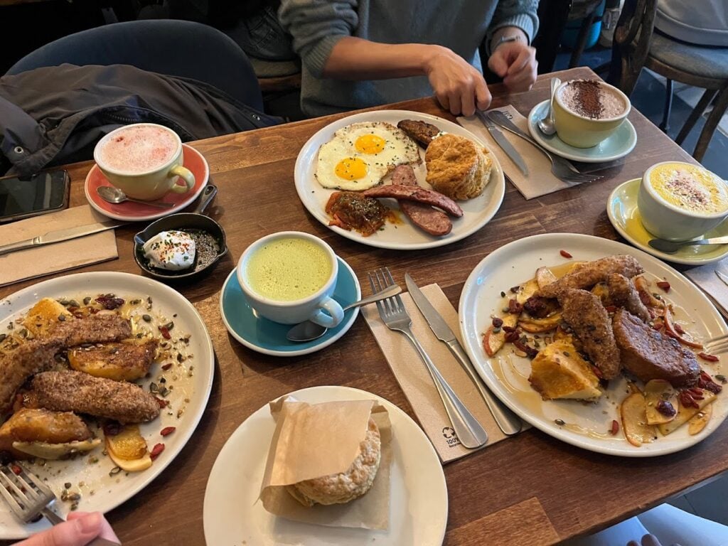 A bustling breakfast scene at Treize Bakery in Paris, with a variety of dishes spread across the table. The feast includes French toast with pomegranate seeds, sunny-side-up eggs with sausages and a scone, a bowl of granola, and beverages like pink cappuccino, green matcha latte, and traditional hot chocolate. This diverse and abundant selection highlights why it's among the best breakfasts in Paris.