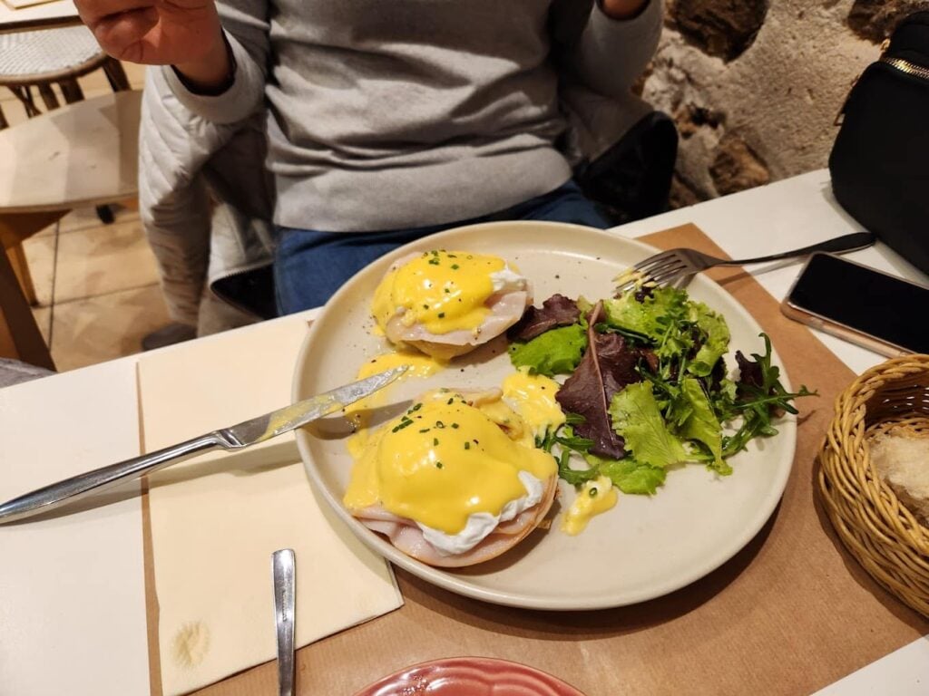 A classic breakfast at Twinkie Breakfast & Lunch in Paris, featuring two perfectly poached eggs on ham and English muffins, generously topped with hollandaise sauce and sprinkled with chives. The dish is served alongside a fresh green salad with mixed leaves and a basket of assorted bread, capturing the essence of what makes it one of the best breakfast in Paris.