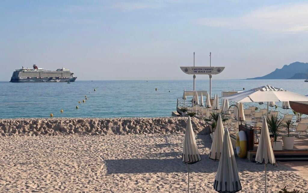 Sandy beach with closed umbrellas and lounge chairs at Carlton Cannes, one of the iconic Cannes beach clubs, with a cruise ship in the calm Mediterranean Sea in the background.