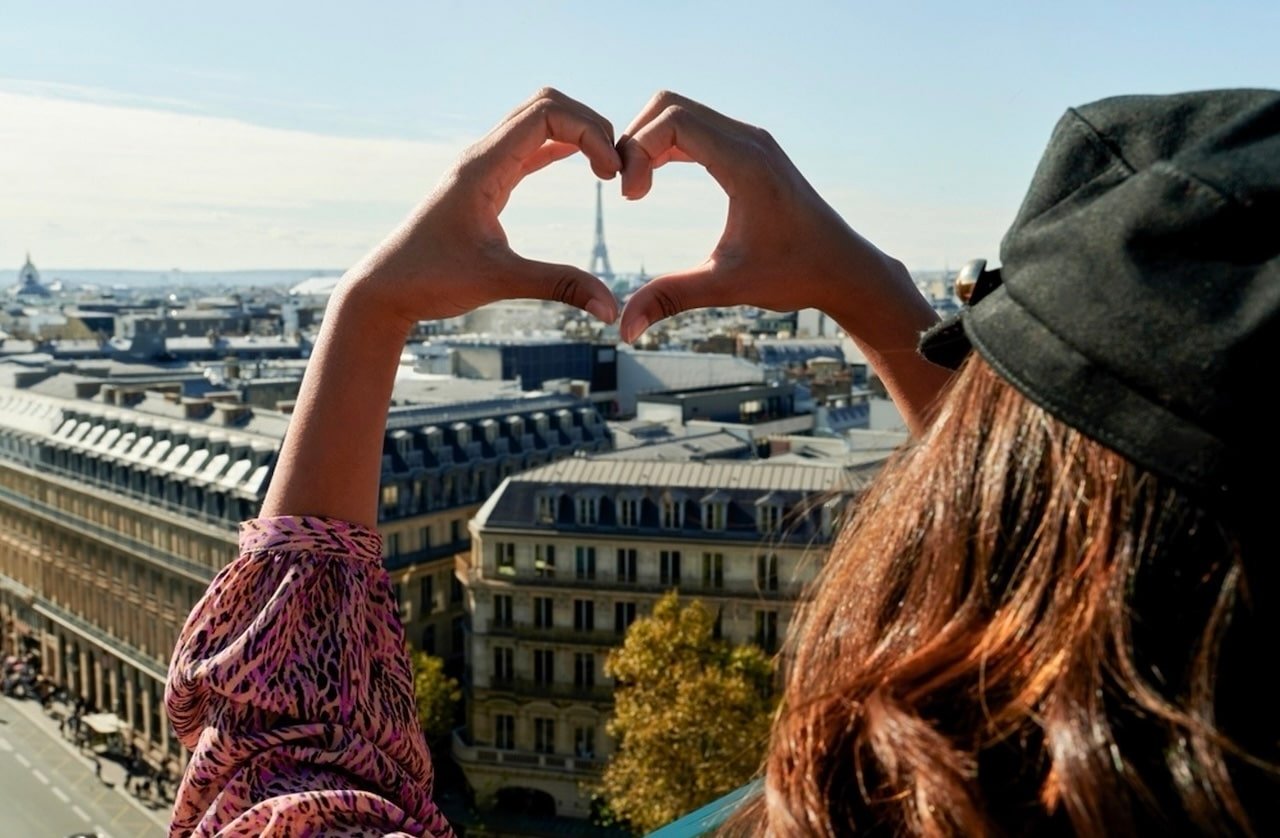 A woman wearing a black beret and a patterned blouse stands on a rooftop overlooking Paris, forming a heart shape with her hands around the distant Eiffel Tower, creating a picturesque scene for a Paris travel blog.
