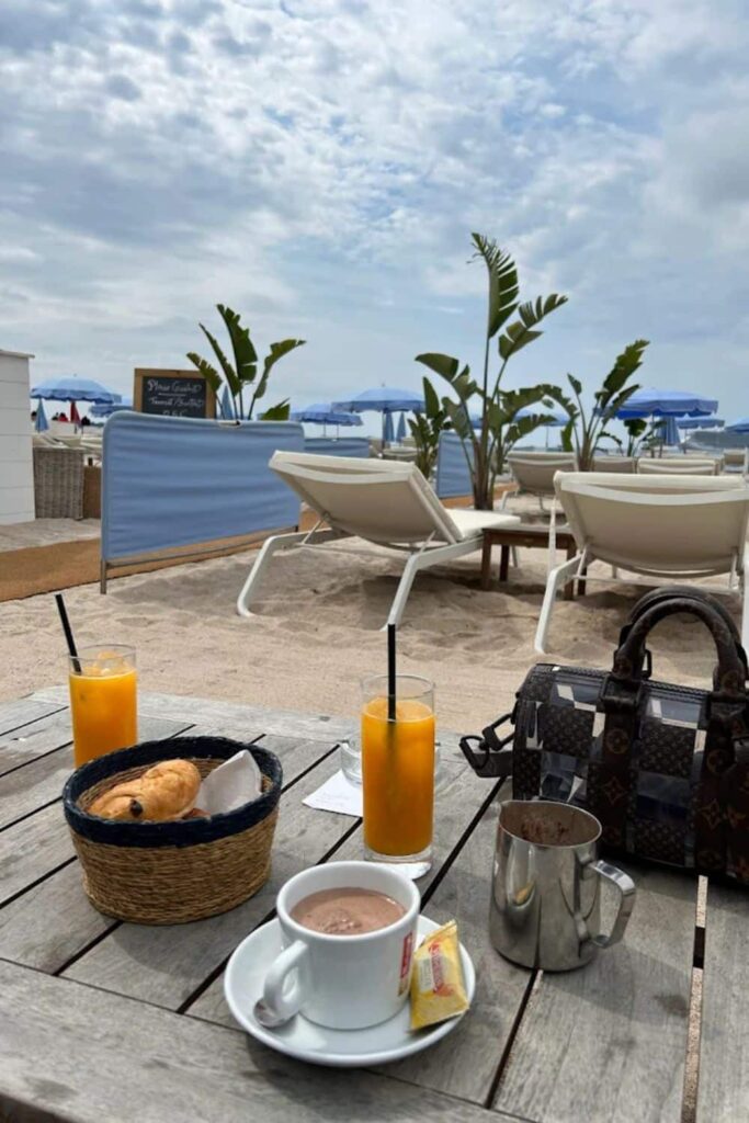 Breakfast setup with croissants, orange juice, and hot chocolate on a wooden table at Plage Goeland, a charming Cannes beach club, with lounge chairs and blue umbrellas in the sandy background.