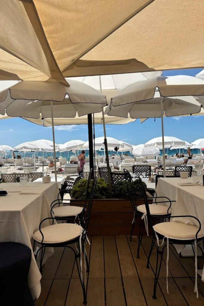 Outdoor dining area with white tablecloths and black wrought-iron chairs overlooking the beach, umbrellas, and sun loungers at one of the popular Cannes beach clubs on a sunny day.